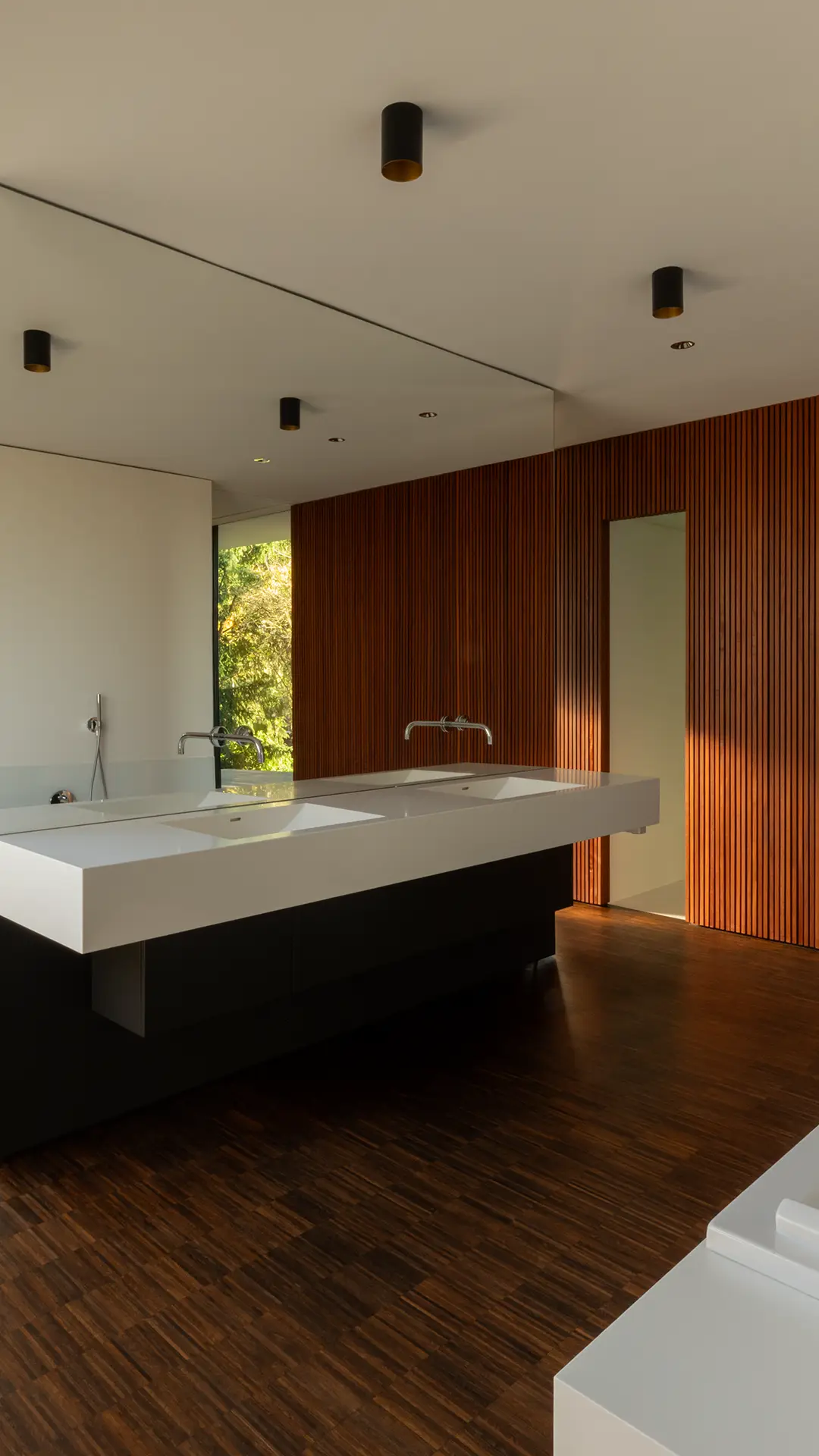 Vertikale Ansicht des Bads im Wohnhaus An Steins Garten in Gießen mit Teaklamellenwand, Mineralwerkstoff-Waschtisch und Badewanne.